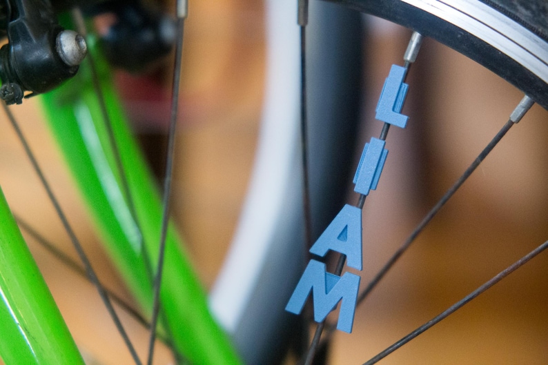 Op de afbeelding: Een close-up van een fietswiel met de naam "LIAM" gespeld in blauwe plastic letters die aan de spaken zijn bevestigd.