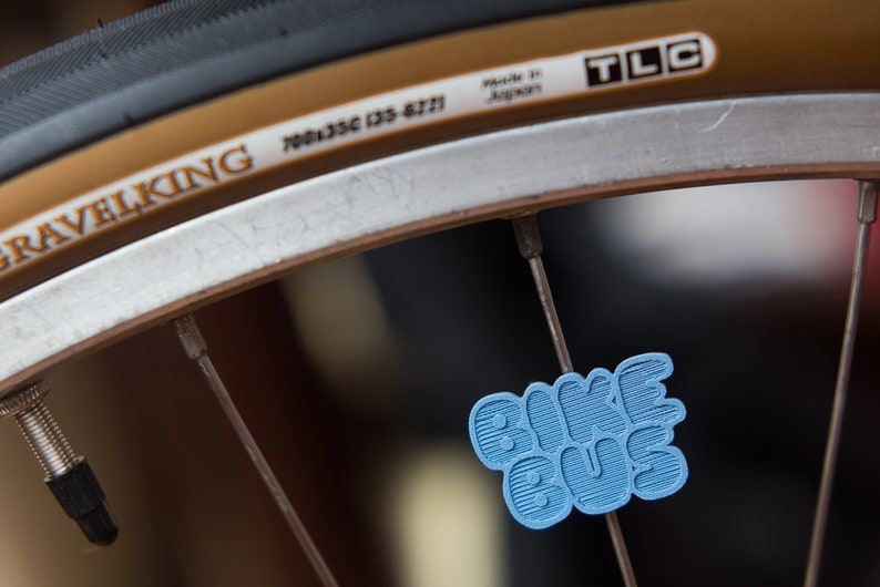 May include: A close-up of a bicycle wheel with a blue Bike Bug sticker attached to a spoke. The wheel has a white rim and a brown tire with the text "Gravelking 700x35C (35-622) Made in Japan TLC" printed on it.
