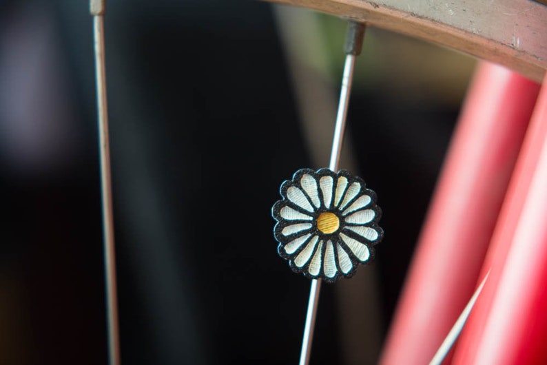 Puede incluir: Un adorno decorativo para radios de bicicleta con forma de margarita. El adorno tiene p&eacute;talos blancos, un borde negro y un centro dorado. Est&aacute; sujeto a un radio plateado, con una llanta roja y plateada en el fondo.