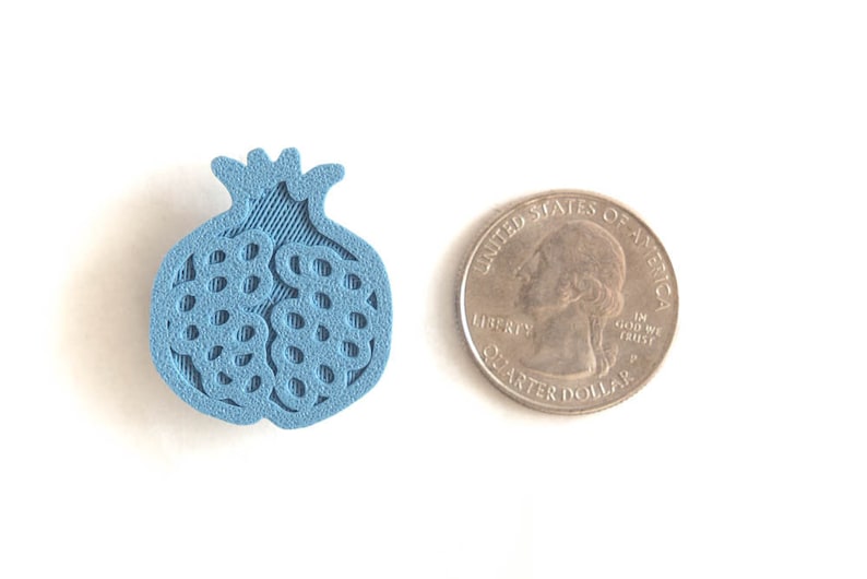 Pu&ograve; includere: Un ciondolo a forma di melograno stampato in 3D di colore azzurro chiaro, circa delle dimensioni di una moneta da 25 centesimi di dollaro americano.