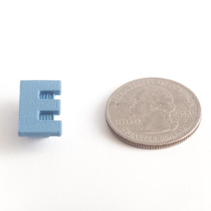 May include: A small, light blue plastic letter E, next to a US quarter for size comparison.
