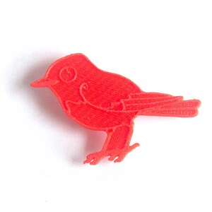 May include: A bright red bird figurine, likely a decorative item or toy, is depicted against a clean white background. The bird has a detailed, textured surface, with visible wings, a beak, and small legs. The overall design is simple and stylized.
