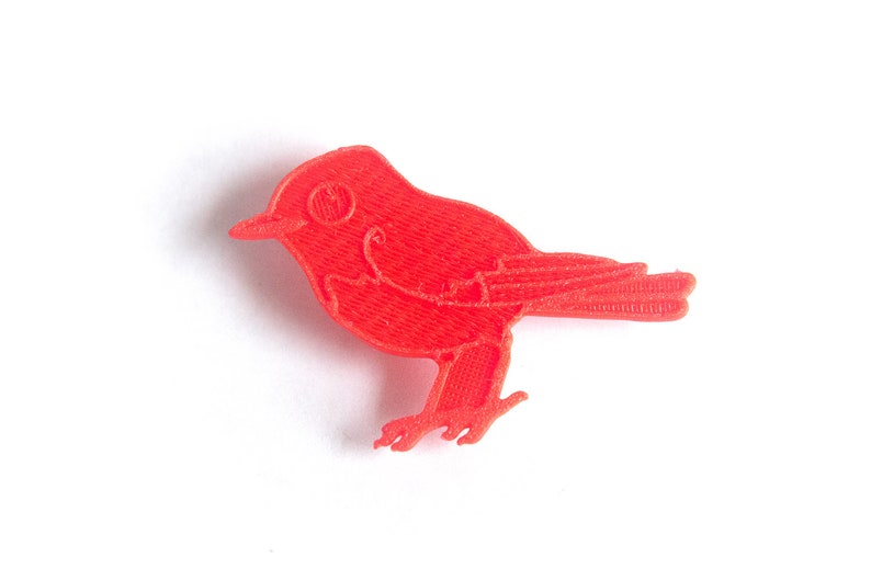 May include: A bright red bird figurine, likely a decorative item or toy, is depicted against a clean white background. The bird has a detailed, textured surface, with visible wings, a beak, and small legs. The overall design is simple and stylized.
