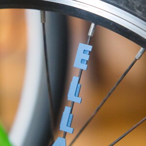 Op de afbeelding: Een close-up van een fietswiel met de naam "Ella" gespeld in blauwe plastic letters die aan de spaken zijn bevestigd.