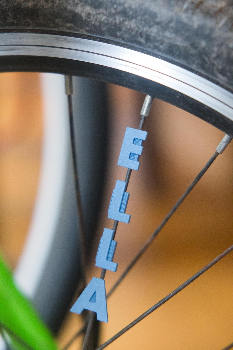 Op de afbeelding: Een close-up van een fietswiel met de naam "Ella" gespeld in blauwe plastic letters die aan de spaken zijn bevestigd.