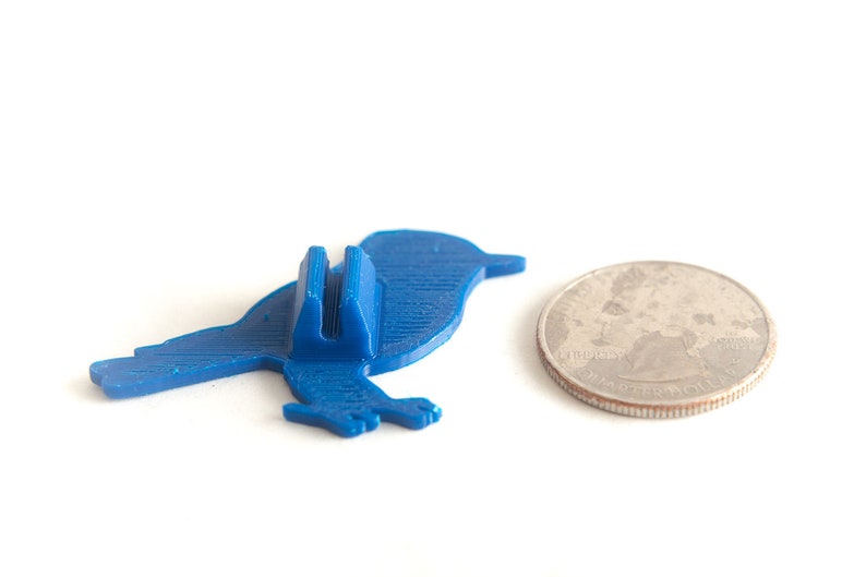 May include: A blue bird-shaped phone stand next to a quarter coin. The stand is made of plastic and has a slot to hold a phone. The quarter is silver and shows a profile of George Washington.