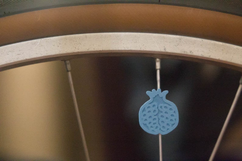 Pu&ograve; includere: Primo piano di una ruota di bicicletta con un ciondolo blu a forma di melograno appeso a un raggio.