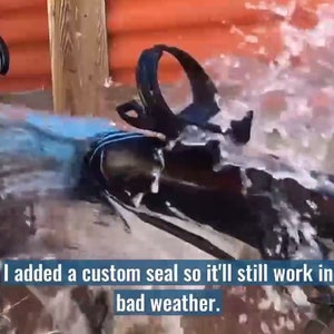 May include: A black bicycle frame with a blue and black seat and a water bottle cage. The bicycle is being sprayed with water. The text "I added a custom seal so it'll still work in bad weather." is visible in the image.