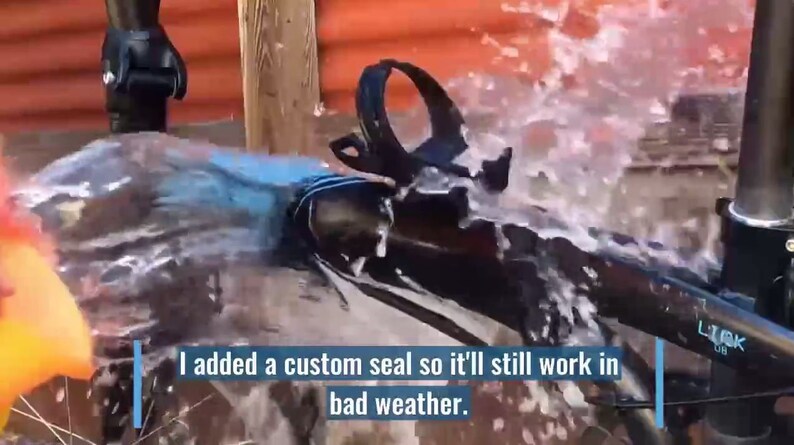 May include: A black bicycle frame with a blue and black seat and a water bottle cage. The bicycle is being sprayed with water. The text "I added a custom seal so it'll still work in bad weather." is visible in the image.