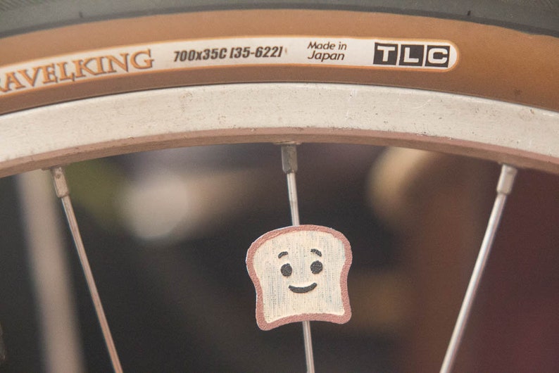Puede incluir: Primer plano de una rueda de bicicleta con un neum&aacute;tico marr&oacute;n y radios plateados. Un peque&ntilde;o amuleto de pan tostado sonriente est&aacute; unido a los radios. El neum&aacute;tico tiene el texto "TRAVELKING" y "Made in Japan".