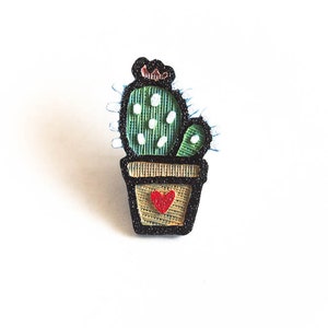 May include: A green cactus with a pink flower on top, in a brown pot with a red heart on the front. The cactus is outlined in black and has white dots on it.