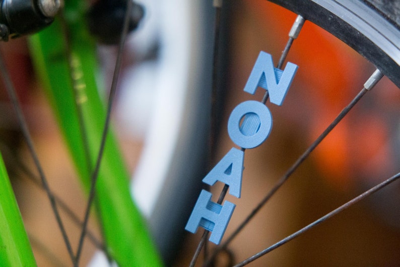 Op de afbeelding: Een fietswiel met de naam "NOAH" gespeld in blauwe letters die aan de spaken zijn bevestigd.