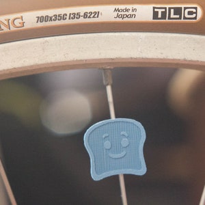 Puede incluir: Primer plano de una rueda de bicicleta con un dije en forma de tostada azul claro sonriente, sujeto a un radio. El neum&aacute;tico tiene el texto "TRAVELKING" y "700x35C (35-622)". Tambi&eacute;n son visibles las palabras "Made in Japan" y "TLC".