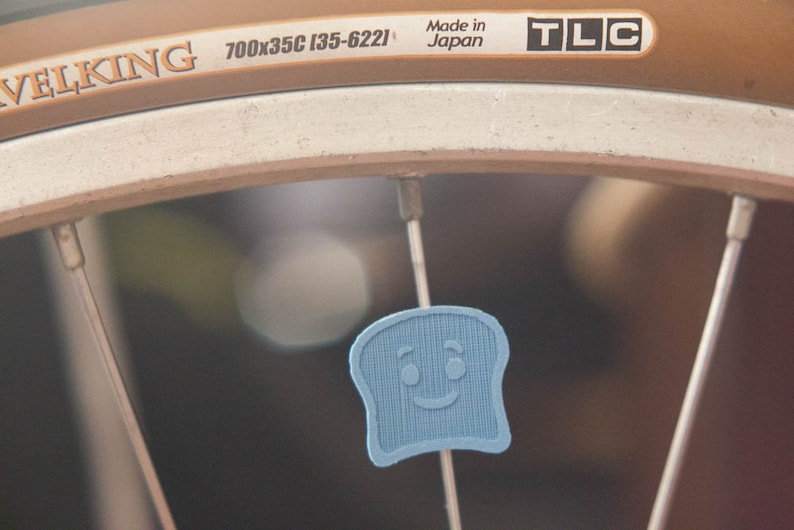 Puede incluir: Primer plano de una rueda de bicicleta con un dije en forma de tostada azul claro sonriente, sujeto a un radio. El neum&aacute;tico tiene el texto "TRAVELKING" y "700x35C (35-622)". Tambi&eacute;n son visibles las palabras "Made in Japan" y "TLC".