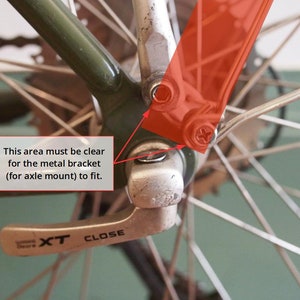 May include: Close-up of a bicycle rear derailleur with a silver lever labelled "XT CLOSE". The derailleur is attached to a green bicycle frame and a silver axle. The area around the axle must be clear for the metal bracket to fit.