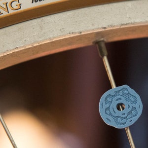Peut inclure: Gros plan sur une roue de v&eacute;lo avec un capuchon de rayon bleu orn&eacute; d'un motif d&eacute;coratif. Le capuchon de rayon est fix&eacute; &agrave; un rayon argent&eacute;.