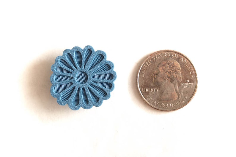 Puede incluir: Un objeto azul claro en forma de flor con un dise&ntilde;o texturizado y detallado, junto a una moneda de veinticinco centavos estadounidense. El dise&ntilde;o del objeto presenta l&iacute;neas radiales y un c&iacute;rculo central.