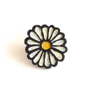Puede incluir: Bot&oacute;n decorativo en forma de margarita con contorno negro, p&eacute;talos blancos y centro amarillo. El bot&oacute;n est&aacute; sobre un fondo blanco liso, resaltando su dise&ntilde;o floral. Adecuado para manualidades y proyectos de costura.