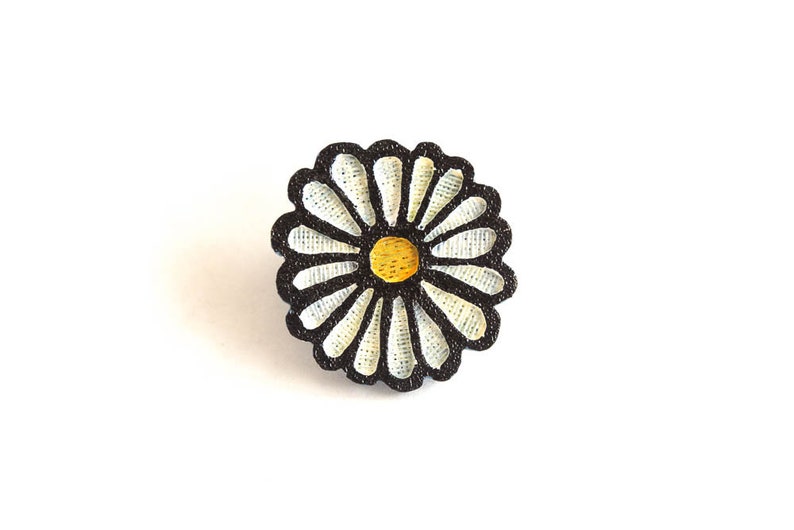Puede incluir: Bot&oacute;n decorativo en forma de margarita con contorno negro, p&eacute;talos blancos y centro amarillo. El bot&oacute;n est&aacute; sobre un fondo blanco liso, resaltando su dise&ntilde;o floral. Adecuado para manualidades y proyectos de costura.