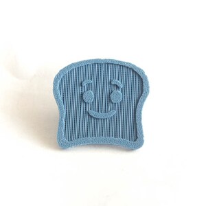 Puede incluir: Pin de rebanada de pan azul claro sonriente. El pin tiene una superficie texturizada con l&iacute;neas verticales y una cara amigable con ojos redondos y una sonrisa curva. Sobre un fondo blanco.