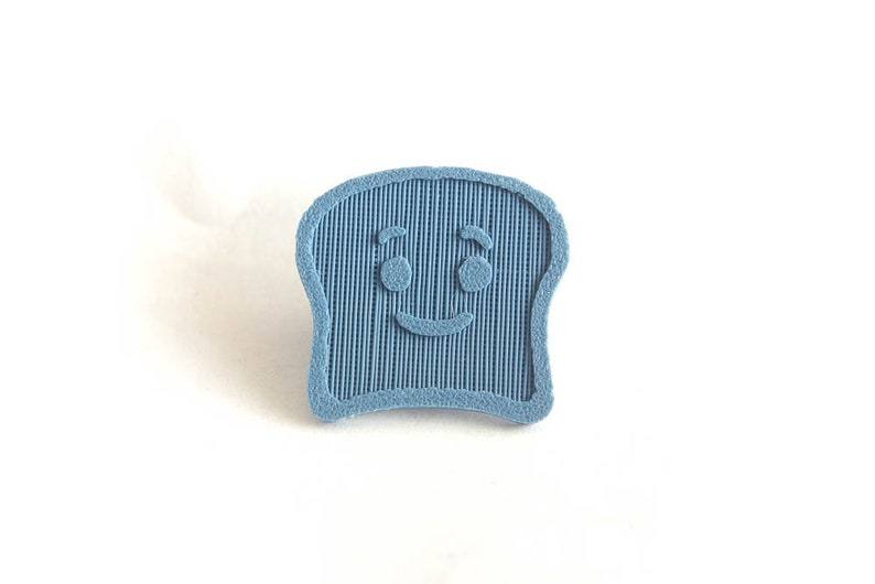 Puede incluir: Pin de rebanada de pan azul claro sonriente. El pin tiene una superficie texturizada con l&iacute;neas verticales y una cara amigable con ojos redondos y una sonrisa curva. Sobre un fondo blanco.