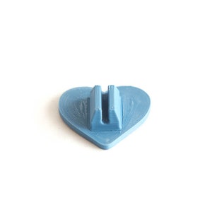 May include: A light blue heart-shaped stand with a slot in the center. The stand is made of plastic and is designed to hold a small object.