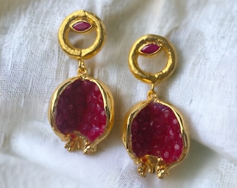 May include: Gold earrings with a red pomegranate design. The earrings feature a small red gemstone in a gold circle.