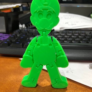 Puede incluir: Una figura de plástico verde de un personaje de la serie de videojuegos Super Mario Bros. La figura está articulada en las articulaciones y tiene una letra "L" en su sombrero.