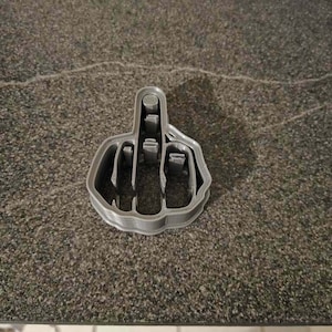 May include: A gray plastic cookie cutter in the shape of a hand giving the middle finger. The cookie cutter is made of a light gray plastic and is sitting on a dark gray surface.