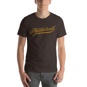 May include: A dark brown t-shirt with a gold-colored "Marblehead" graphic in a cursive font.
