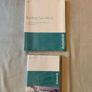 May include: Two Autodesk software manuals. The top manual is white and teal, titled Building Your World, Conceptual Design and Visualization with AutoCAD 2003. The bottom manual is white and teal, titled AutoCAD 2007, featuring a technical image.