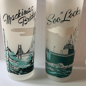 May include: Two frosted glass tumblers with black and teal illustrations. One features the Mackinac Bridge, the other the Soo Locks, both with corresponding text. The glasses have a vintage aesthetic and are likely souvenir items.