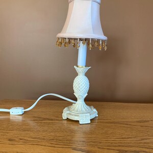 May include: A table lamp featuring a white pineapple-shaped base with a textured surface and a light pink fabric lampshade. The shade is decorated with a fringe of small, amber-colored, teardrop-shaped beads hanging from the bottom edge.