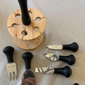 May include: A cheese serving set featuring five stainless steel utensils with black handles, each marked GourmetBuffet. The set includes a fork, a spade, and three knives of varying shapes, accompanied by a round wooden storage block.