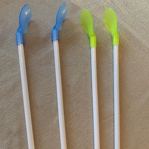 May include: Four plastic stir sticks with paddle-shaped ends. Two have blue paddles and two have green paddles. The sticks are white with blue or green accents. The items are arranged on a neutral-colored surface.