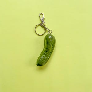 May include: A green pickle keychain with a silver-colored clasp and chain. The pickle has a textured surface and is suspended against a yellow-green background. The keychain is a fun, novelty item.