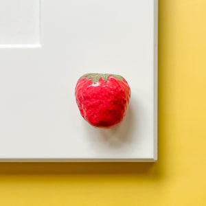 May include: A red ceramic strawberry cabinet knob with a green stem. The knob is mounted on a white surface, with a yellow background. This decorative knob adds a touch of whimsy to any cabinet or drawer.