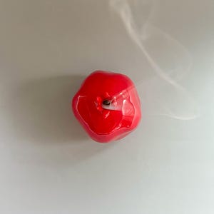 Op de afbeelding: Een rode, appelvormige kaars met een afgebrande lont. De kaars is glanzend en heeft een glad oppervlak. Wispelturige rook stijgt op van de lont, wat aangeeft dat deze recentelijk is gedoofd.