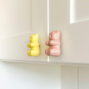 May include: Two gummy bear-shaped cabinet knobs. One is yellow, the other is pink. They are mounted on a white cabinet door. The knobs are made of ceramic and have a glossy finish. The hardware is gold.