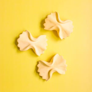 May include: Three uncooked farfalle pasta shapes on a yellow background.