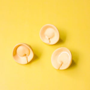 May include: Three beige, ear-shaped ceramic objects are arranged on a bright yellow background. Each object has a curved, rounded form with a small indentation. The objects are evenly spaced, creating a visually appealing composition.
