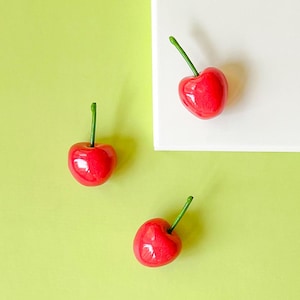 Puede incluir: Tres cerezas rojas brillantes con tallos verdes se disponen sobre un fondo verde claro y blanco. Las cerezas tienen un acabado brillante, lo que sugiere que son artificiales o estilizadas.