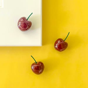 Op de afbeelding: Drie glanzende, donkerrode kersenornamenten met groene stelen zijn gerangschikt tegen een heldergele achtergrond en een witte vierkant. De kersen hebben een realistische uitstraling, wat duidt op een fruit-thema decoratie.