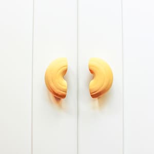May include: Two cabinet door handles with a curved, cashew-like design. The handles are a warm yellow colour with a gradient of brown at the edges, set against a white background. The handles are symmetrical and appear to be made of wood.