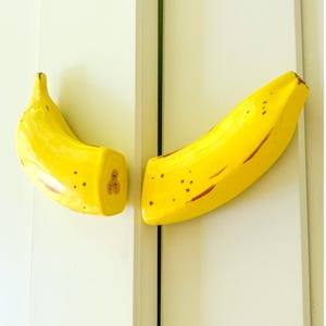 May include: Two yellow banana-shaped cabinet pulls. The pulls are painted with brown spots and streaks, and one is cut to reveal the interior. These decorative pulls add a playful touch to any cabinet or drawer.