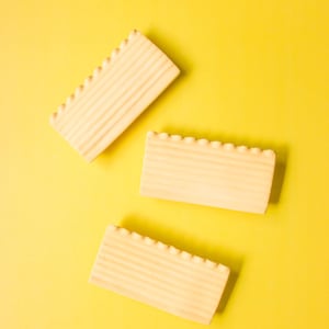 Könnte beinhalten: Drei rechteckige, hellfarbene Holzblöcke mit geriffelten Kanten und gerillten Oberflächen sind auf einem leuchtend gelben Hintergrund angeordnet. Die Blöcke sind in verschiedenen Winkeln positioniert, was eine ansprechende Komposition ergibt.