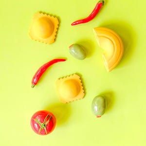 Puede incluir: Una vibrante disposición de piezas de cerámica con temática de alimentos sobre un fondo verde brillante. La composición incluye ravioles amarillos, chiles rojos, aceitunas verdes, un tomate rojo y una gran forma de pasta amarilla.