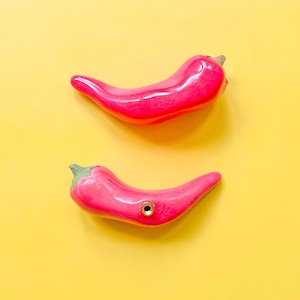 May include: Two bright pink ceramic chilli peppers on a yellow background. One pepper has a small hole with a gold-coloured ring. The peppers are glossy and have a realistic shape, suggesting a kitchen or food-related theme.