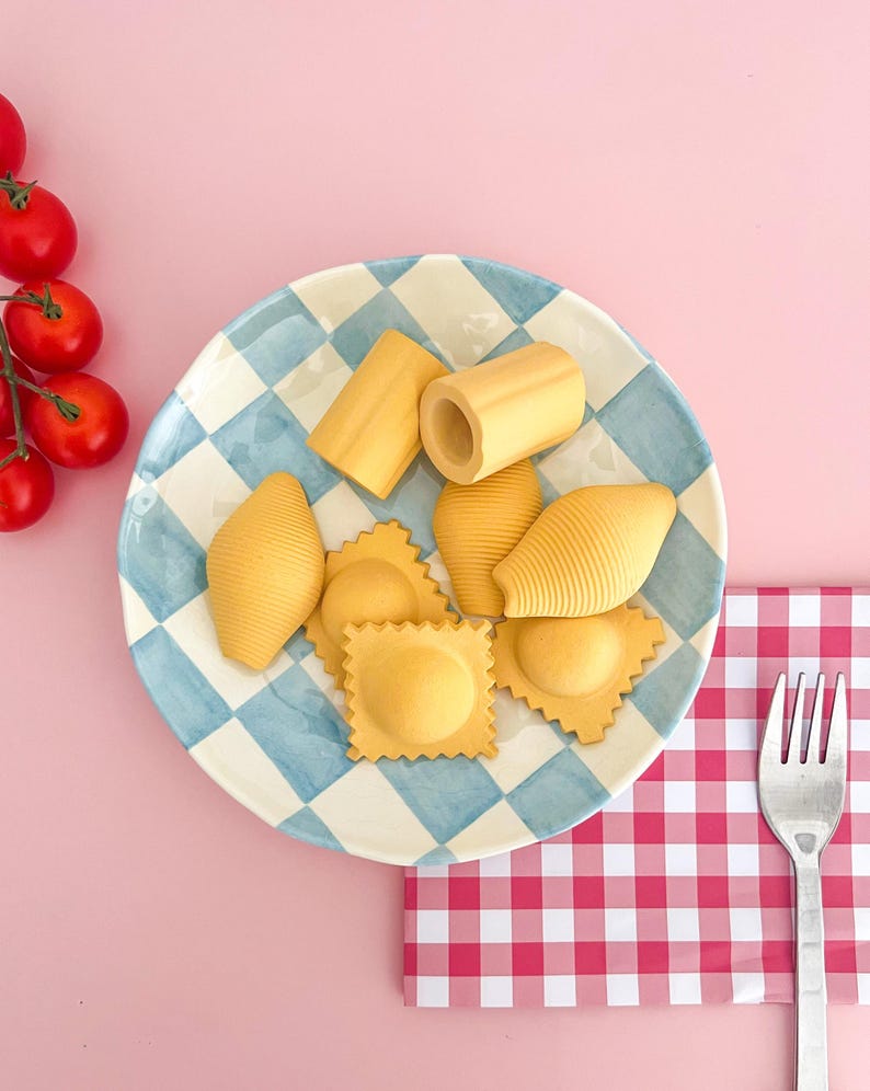 May include: A blue and white checked plate with various types of pasta, including ravioli, shells, and tubes. The plate is on a pink surface with a red and white checked napkin and a silver fork.