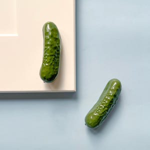 May include: Two green pickle-shaped cabinet knobs. One is attached to a cream-coloured cabinet door, and the other is lying on a light blue surface. The knobs have a textured surface and a glossy finish.